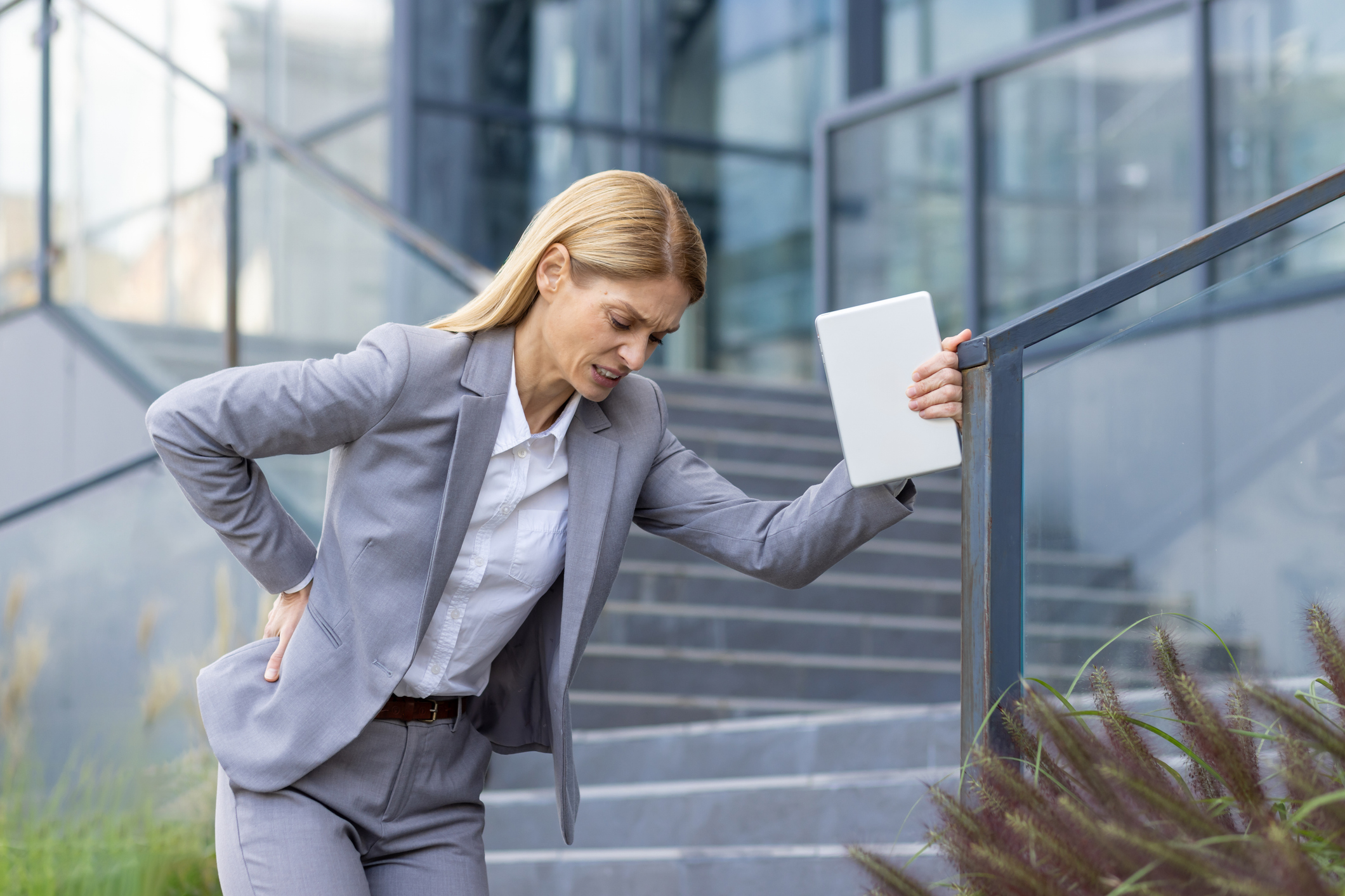 Tired young business woman standing near the office building, leaning her hands on the stair railing and holding her back, feeling severe pain